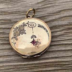 Peut inclure: Un médaillon en or avec un motif floral et une petite pierre rose au centre. Le médaillon a un couvercle à charnière et une boucle pour attacher une chaîne.