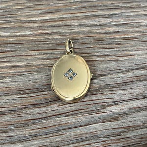 Könnte beinhalten: Ein ovaler Goldmedaillon-Anhänger mit einer glatten, polierten Oberfläche und einer kleinen, mittig platzierten quadratischen Gruppe aus vier winzigen, klaren, funkelnden Steinen. Der Anhänger hat eine integrierte Goldöse zur Befestigung.