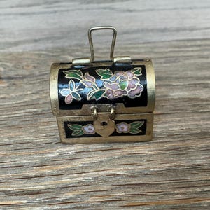 Puede incluir: Pequeño cofre negro y dorado con diseños florales en rosa, verde y blanco. El cofre en miniatura tiene una tapa con bisagras y un asa de metal. El cofre está sobre una superficie de madera.