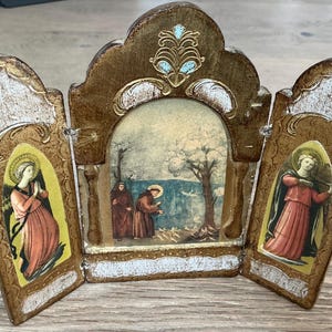 Puede incluir: Un icono religioso vintage de tres paneles con marco dorado e imágenes pintadas. El panel central representa figuras cerca de una masa de agua, mientras que los paneles laterales muestran santos con túnicas rojas y amarillas. El icono tiene una apariencia envejecida y antigua.