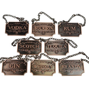 May include: Set of seven antique gold bottle tags with black chains. The tags are engraved with the names of different types of alcohol: Vodka, Bourbon, Rum, Scotch, Tequila, Gin, Whiskey, and Brandy.
