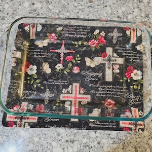 May include: A rectangular clear glass baking dish sits atop a black fabric mat featuring a repeating pattern of red and white roses, butterflies, crosses, and the text Amazing Grace in a script font, with a gold-toned metal utensil visible.