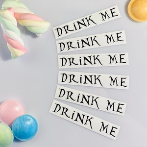 May include: Six white rectangular labels with the text "Drink Me" printed in black. The labels are arranged in a vertical stack, with the top label slightly offset to the right.