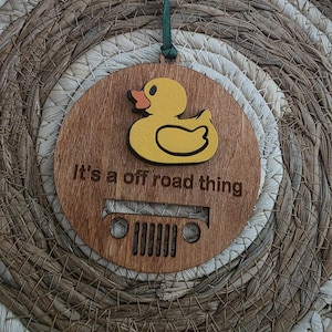 May include: A wooden ornament with a yellow rubber ducky and the text "It's a off road thing." The ornament has a Jeep grill design and a green ribbon for hanging. The background is a woven, natural-toned material.