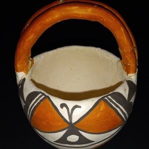May include: A handmade, small, off-white ceramic basket with a curved orange handle. The basket's exterior features a geometric design in orange, black, and white. The design includes a butterfly-like shape. The background is black.