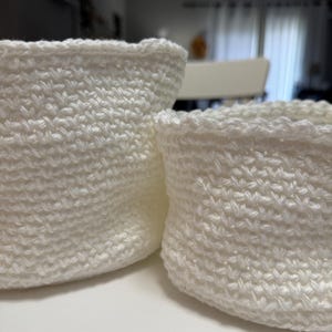 May include: Two small, off-white crochet baskets. The baskets are cylindrical with a slightly flared top and a textured stitch pattern. The crochet work appears tight and even, suggesting a handmade item. The baskets are likely for storage or decorative purposes.