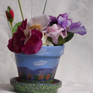 Puede incluir: Una maceta de terracota pintada de azul con una escena de paisaje y nubes blancas. La maceta tiene un platillo a juego y está llena de una variedad de flores artificiales, incluyendo rosas, violetas y blancas.