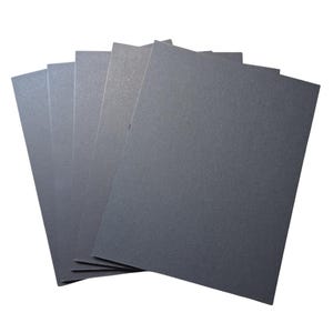 May include: A stack of gray sandpaper sheets fanned out against a white background. The sandpaper has a fine, textured surface, and the sheets are rectangular in shape. The image is well-lit, showing the details of the sandpaper.