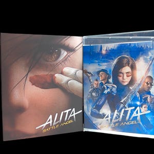 May include: A Blu-ray movie case for "Alita: Battle Angel" is open, revealing the disc and cover art. The cover art features a woman with a sword and other characters in a futuristic setting. The case on the left shows a close-up of a face with the movie title.