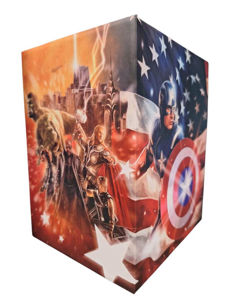 K&ouml;nnte beinhalten: W&uuml;rfelf&ouml;rmige Box mit einer Collage von Marvel-Superhelden. Die Illustration zeigt Captain America, Thor und Hulk. Der Hintergrund zeigt eine amerikanische Flagge und eine Stadtlandschaft mit Blitzen.