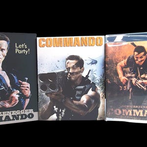 May include: Three movie cases for the film "Commando" starring Arnold Schwarzenegger. The cases feature images of Schwarzenegger holding weapons and the film's title. One case has the text "Let's Party!"