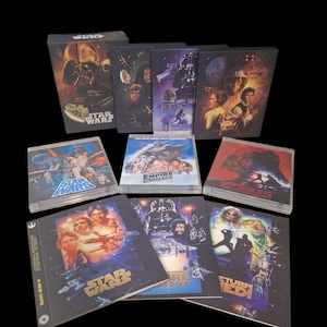 May include: A collection of Star Wars movie box sets and cases, featuring iconic characters and scenes. The cases display movie titles like "A New Hope", "Empire Strikes Back", and "Return of the Jedi". The artwork showcases characters like Darth Vader and Luke Skywalker.