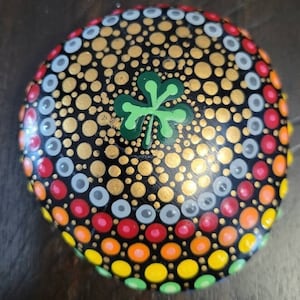 May include: A round, black stone painted with a mandala design. The center of the mandala features a green four-leaf clover. The outer edges of the mandala are painted in a rainbow of colors, including red, orange, yellow, green, blue, and purple.