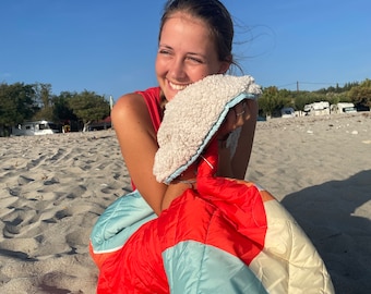 May include: A colorful, quilted blanket with red, blue, and cream sections. The blanket has a soft, fluffy white and blue trim. The person is holding the blanket on a sandy beach under a clear blue sky.
