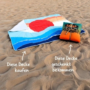 Puede incluir: Una manta de playa con un diseño de puesta de sol, con un gran sol rojo y olas azules. Una almohada a juego está atada con hilo. La manta está doblada en una playa de arena.