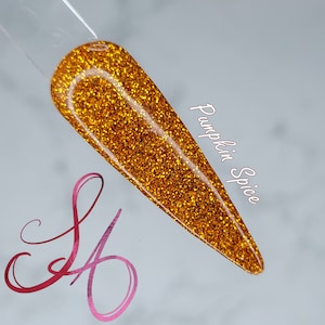 Puede incluir: Primer plano de un esmalte de uñas de purpurina dorado con el texto "Pumpkin Spice" escrito en el lado derecho de la imagen.