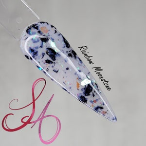 May include: A white nail polish with black and iridescent glitter. The polish is called "Rainbow Moonstone".