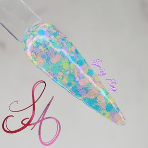 Puede incluir: Primer plano de un esmalte de uñas azul claro con una variedad de purpurina de colores pastel. El texto "Spring Fling" está escrito en morado en el lado derecho de la imagen.