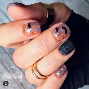 May include: Close-up of a hand with several fingernails painted with a Halloween theme.  One nail is painted black with a textured finish.  Another nail is painted with a light purple and orange ombre effect with black bats and gold glitter.  The text "The.last.Nail" is visible in the lower left corner of the image.