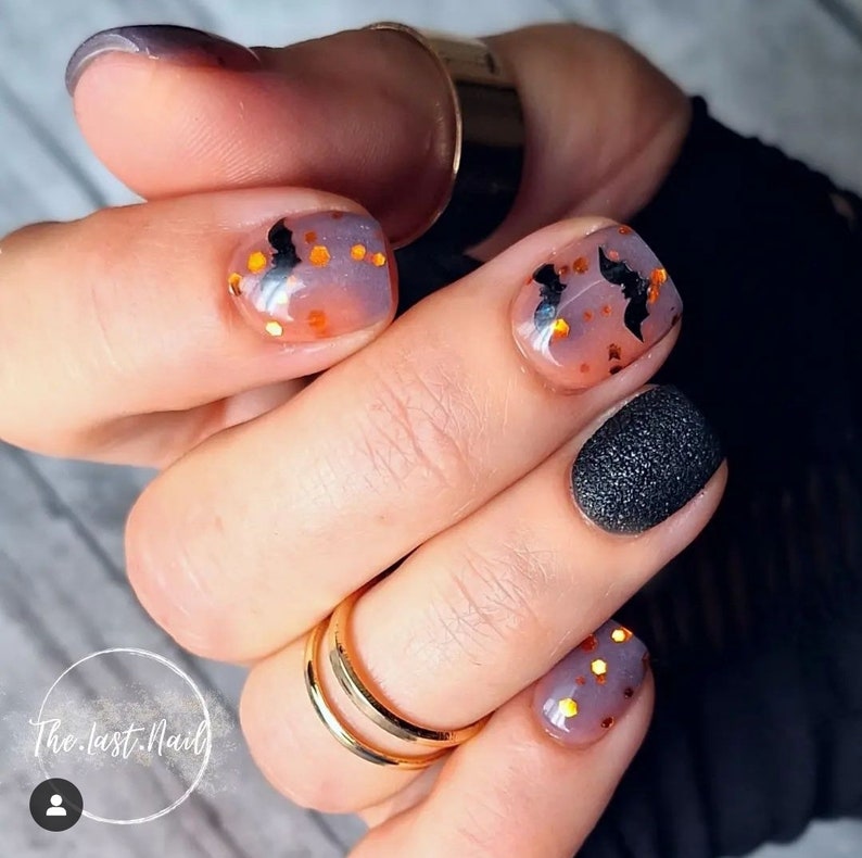 May include: Close-up of a hand with several fingernails painted with a Halloween theme.  One nail is painted black with a textured finish.  Another nail is painted with a light purple and orange ombre effect with black bats and gold glitter.  The text "The.last.Nail" is visible in the lower left corner of the image.