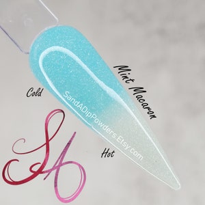 Puede incluir: Un polvo de inmersión para uñas transparente que pasa de un azul claro a un blanco. El polvo está etiquetado como "Mint Macaron" y "SandADipPowders Etsy.com". Las palabras "Frío" y "Caliente" están escritas a ambos lados del polvo.