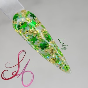 May include: A clear nail with green shamrock and gold foil accents. The nail is decorated with glitter and the word "Lucky" in green script. The nail is on a white background.