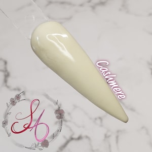 Può includere: Un campione di unghia con un colore bianco sporco cremoso, etichettato "Cashmere". L'unghia è lunga e appuntita, esposta su un bastoncino di plastica trasparente. Lo sfondo è un motivo marmorizzato con un logo floreale.