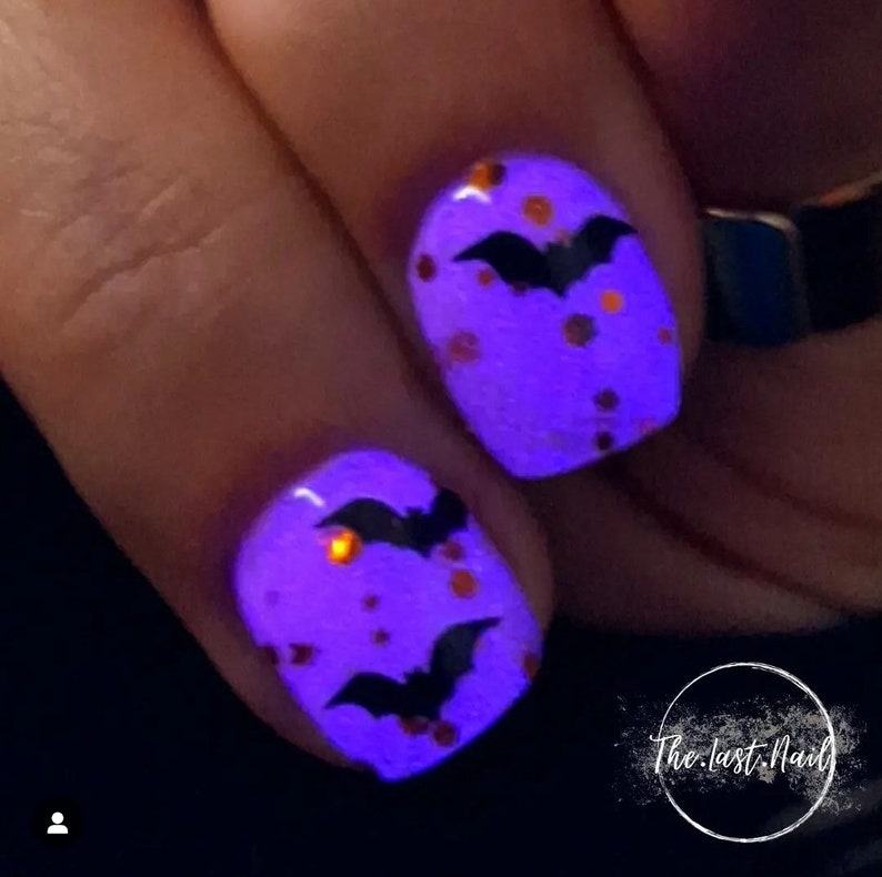 May include: Two fingernails painted a bright purple with black bat silhouettes and orange glitter. The nails are glowing under a black light.