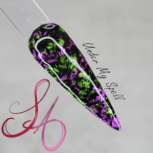 May include: A black nail polish with green and purple iridescent glitter flakes. The text "Under My Spell" is written on the side of the nail.