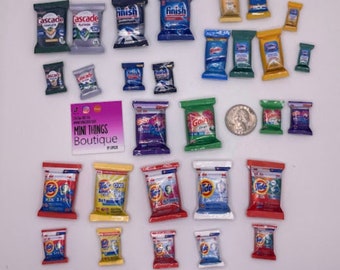 May include: A collection of miniature laundry detergent packets in various colors and brands, including Tide, Gain, Cascade, Finish, and OxiClean. The packets are arranged in rows on a white surface. The text "Mini Things Boutique" is visible on a small sign.