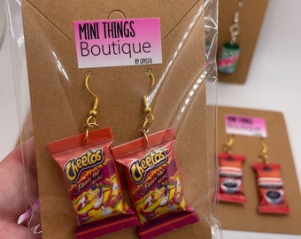May include: A pair of gold hoop earrings with red and yellow Cheetos Flamin' Hot snack bags hanging from them. The earrings are displayed on a brown card with the text "MINI THINGS Boutique by CRYSTAL".