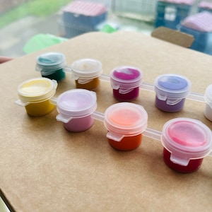 May include: A set of ten small paint pots with lids, arranged in a row on a brown surface. The pots are filled with different colours of paint, including yellow, orange, red, pink, purple, blue, green, and brown.