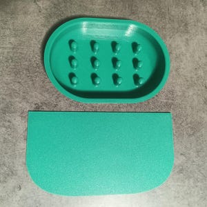 Puede incluir: Un jabonera ovalada de color verde azulado con múltiples agujeros pequeños en la parte inferior. La jabonera está hecha de plástico liso y duro. También se incluye una jabonera rectangular verde azulado a juego.