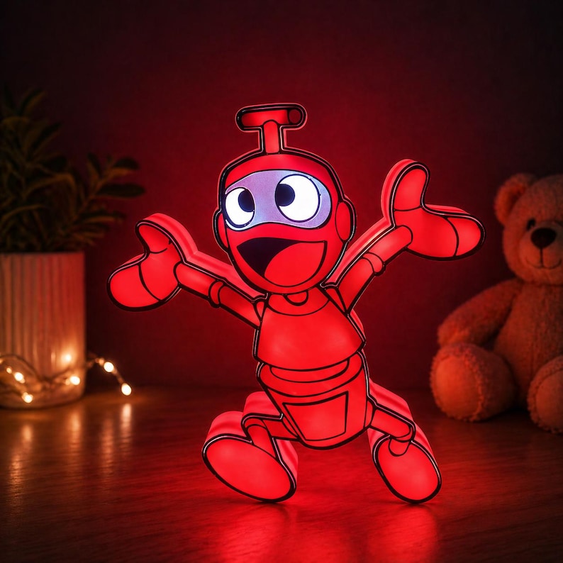 May include: A red robot-shaped LED night light with a happy expression, arms outstretched, and a blue visor. The light is on a wooden surface, with a teddy bear and a plant in the background.