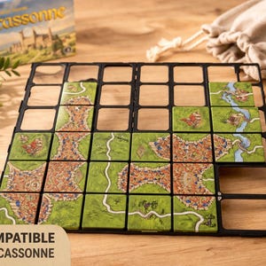 Könnte beinhalten: Ein Brettspiel-Set mit einem schwarzen Raster, grünen und braunen Kacheln und einer Schachtel mit der Aufschrift "Carcassonne". Die Kacheln zeigen Illustrationen von Straßen, Gebäuden und Feldern. Der Text "COMPATIBLE CARCASSONNE" ist sichtbar. Ein beiger Kordelzugbeutel ist im Hintergrund.