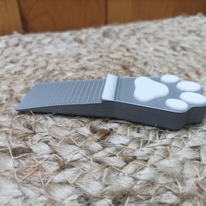 May include: A grey doorstop shaped like a paw print with white paw pads. The doorstop is made of a textured material and has a slight incline.