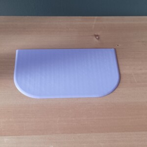 May include: A light purple plastic scraper with a rounded edge, sitting on a wooden surface.
