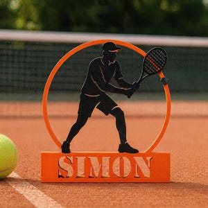 Puede incluir: Decoración naranja con temática de tenis que presenta la silueta de un tenista dentro de un círculo naranja. La base de la decoración tiene el nombre "SIMON" recortado. Una pelota de tenis está en la cancha.