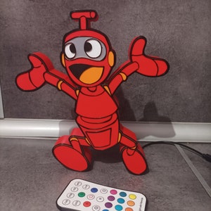 May include: A red robot-shaped LED lamp with a happy expression and outstretched arms. The robot has black outlines, orange accents, and a gray face. A remote control with color options is in front of the lamp.