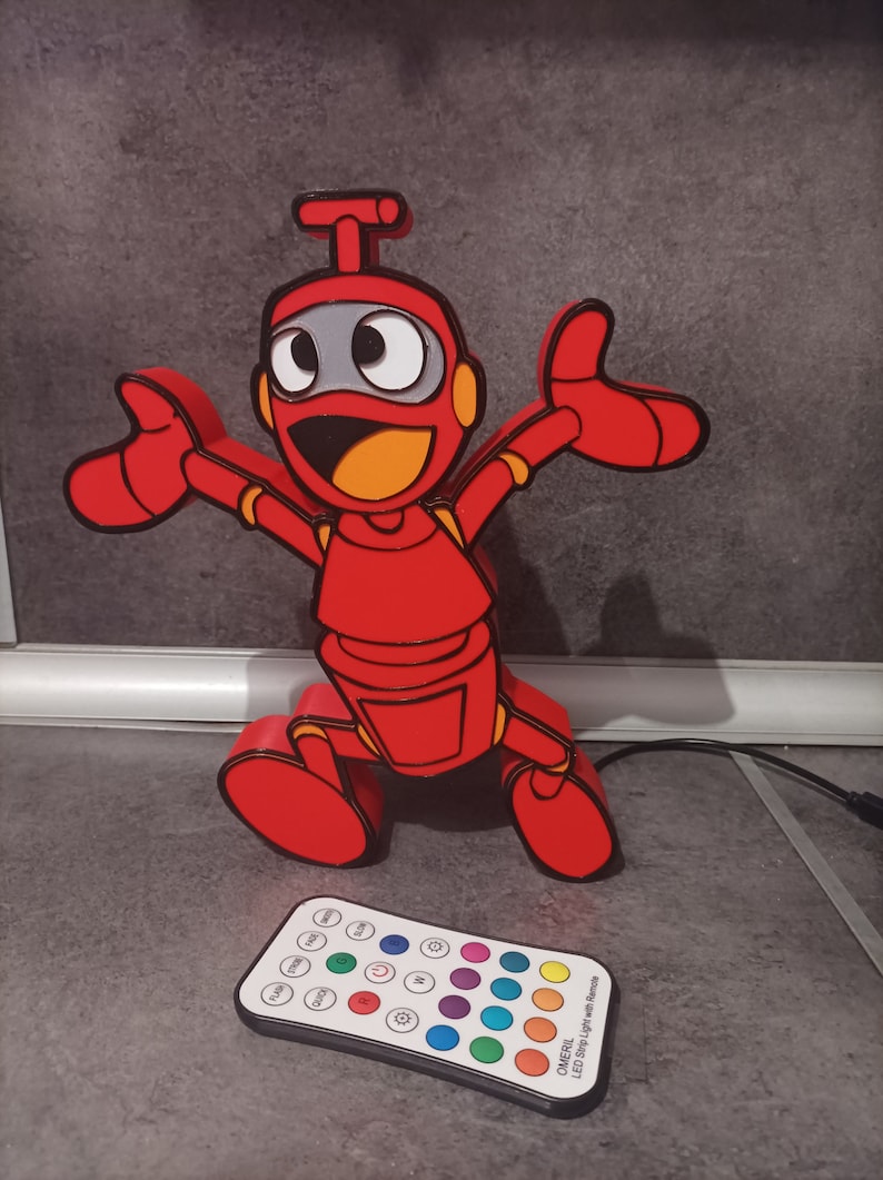 May include: A red robot-shaped LED lamp with a happy expression and outstretched arms. The robot has black outlines, orange accents, and a gray face. A remote control with color options is in front of the lamp.