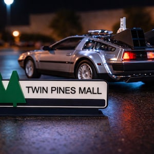 May include: A silver DeLorean model car with a black rear section and red tail lights is parked near a sign that reads "TWIN PINES MALL". The sign has two green tree silhouettes. The scene is set at night.