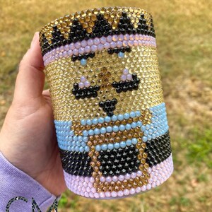 May include: A gold, black, blue, and pink rhinestone tumbler with a cartoon face design. The face has a mustache, a hat, and a cup of coffee.