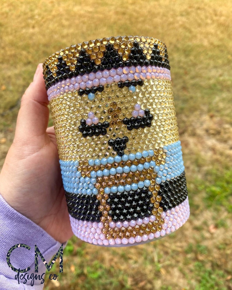 May include: A gold, black, blue, and pink rhinestone tumbler with a cartoon face design. The face has a mustache, a hat, and a cup of coffee.