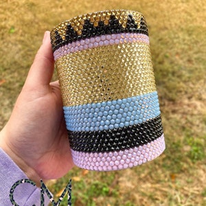 May include: A gold, light blue, black, and pink tumbler covered in rhinestones.