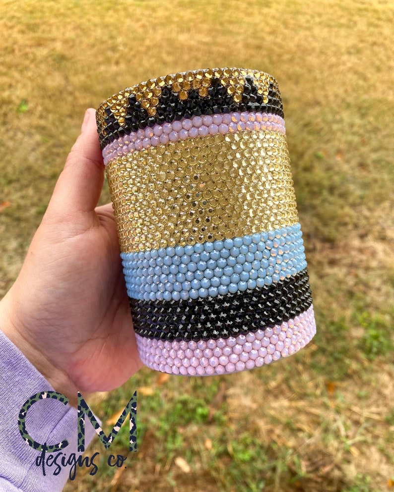May include: A gold, light blue, black, and pink tumbler covered in rhinestones.