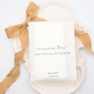 May include: White tissue paper envelope with the text "To catch the tears that love has left behind" and "John Smith 1954-2024". The envelope is on a white plate with a gold ribbon.