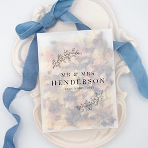 May include: A translucent packet filled with colorful flower petals, with the text "MR & MRS HENDERSON 12TH MARCH 2026" in black. The packet is decorated with floral illustrations and sits on a white, ornate plate, with a blue ribbon.