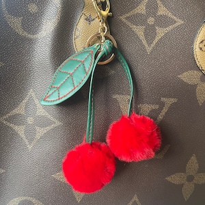 May include: A green leather leaf charm with red fuzzy pom-poms attached to a gold chain. The charm is attached to a brown leather bag.