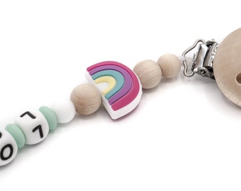 Può includere: Un mordedor in silicone arcobaleno con un perno di legno e una clip. Il mordedor ha un design arcobaleno con colori rosa, viola, giallo e verde. Il perno di legno è in legno naturale. La clip è in metallo argentato.