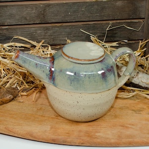 Könnte beinhalten: Eine handgefertigte Keramik-Teekanne mit einem gesprenkelten cremefarbenen Boden und einer hellblau und violett glasierten Oberseite. Die Teekanne hat einen passenden Deckel und einen gebogenen Griff. Der Ausguss ist orange-rot.